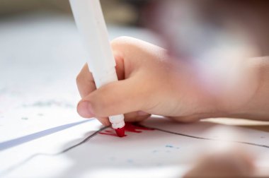 Elinde kırmızı kalemle kağıt üzerine çizen küçük bir çocuğun eli ve ön planda odaklanamadığı şey gözlüğü ve kafası.
