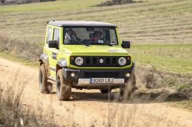 Madrid, İspanya; 02-15-2025: Yeşil bir Suzuki Jimny off-road rallisinde toz ve çamuru tekmeleyerek toprak bir pistte hızla ilerliyor. Yardımcı sürücü ve şoför kask takıyor, yarışa odaklanıyor.