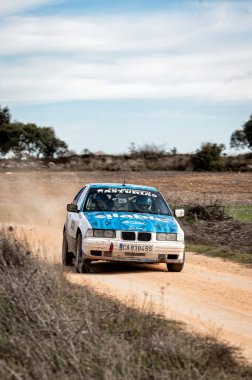 Madrid, İspanya; 02-15-2025: Mavi ve beyaz bir BMW 323i E36 bir çakıl rali pistinde geziniyor ve kırsal alanda hızla toz kaldırıyor. Yardımcı sürücü ve şoför ileriye odaklanıyor.
