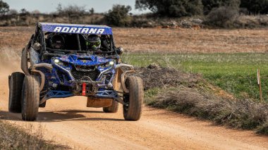 Madrid, İspanya; 02-15-2025: Mavi bir CAN-AM Maverick X3 tozlu bir off-road pistinde hızla ilerliyor, engebeli arazide tekerleklerini kaldırıyor. Şoför ve yardımcı sürücü koruma için kask takıyor.