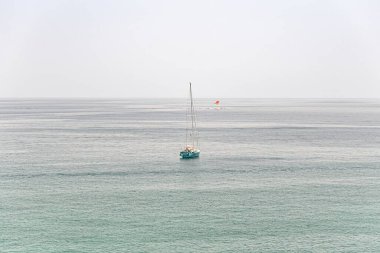 Açık ufukta turkuaz bir denizde yüzen yelkenli. Sisli bir gökyüzünün altında renkli paraşütlü bir paraşüt.