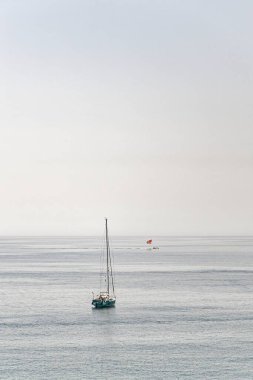 Açık ufukta turkuaz bir denizde yüzen yelkenli. Sisli bir gökyüzünün altında renkli paraşütlü bir paraşüt.