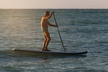 Emekli adam şafakta sörf tahtasıyla denizde yüzüyor.