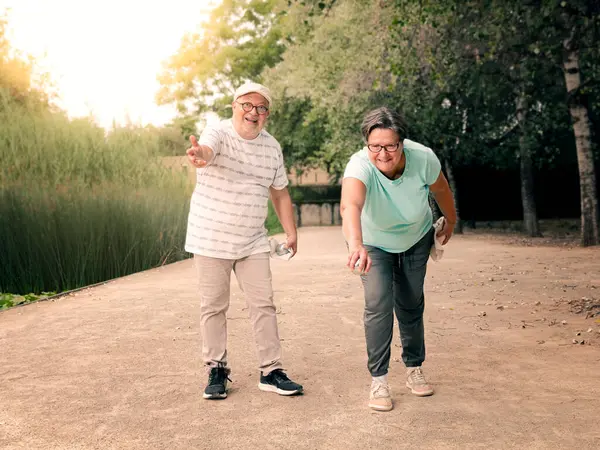 Şehir bahçesinde Petanque oynayan bir çift. Arka planda gölet olan adam atışları izliyor..