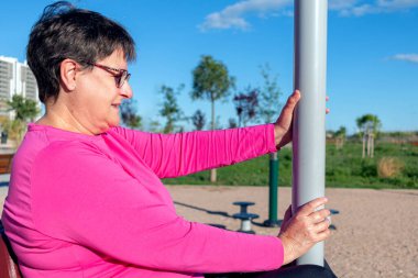 Halka açık bir parkta egzersiz makinesinde oturan yaşlı bir kadının orta boy portresi..