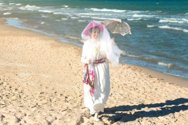 Genç bir kadın, Joaqun Sorolla 'nın Clotilde tablosunun şemsiyesi ve şapka uyarlaması ile sahilin kıyısında gezinir.