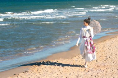 Genç bir kadın, Joaqun Sorolla 'nın Clotilde tablosunun şemsiyesi ve şapka uyarlaması ile sahilin kıyısında gezinir.