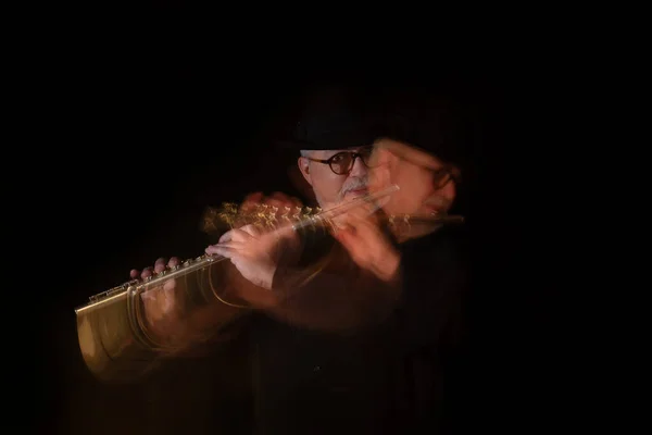 Senior adult with white beard and round glasses making music on music day with handmade Egyptian flute decorated with inlay in front of black piano with hat. Black background. Low key