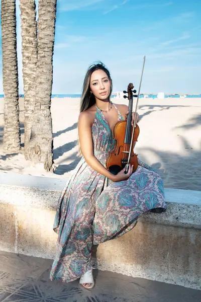 woman violinist sitting on a beach promenade with the sea in the background looking at the camera. Sunny afternoon among palm trees