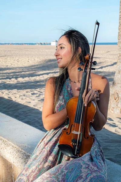 woman violinist sitting on the beach with her violin looking to her right off camera. Sunny afternoon by a palm tree