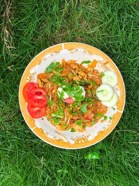 Dilimlenmiş salatalık, domates ve kereviz yaprakları gibi çeşitli garnitürlerle servis edilen bir tabak Tiaw Noodles.