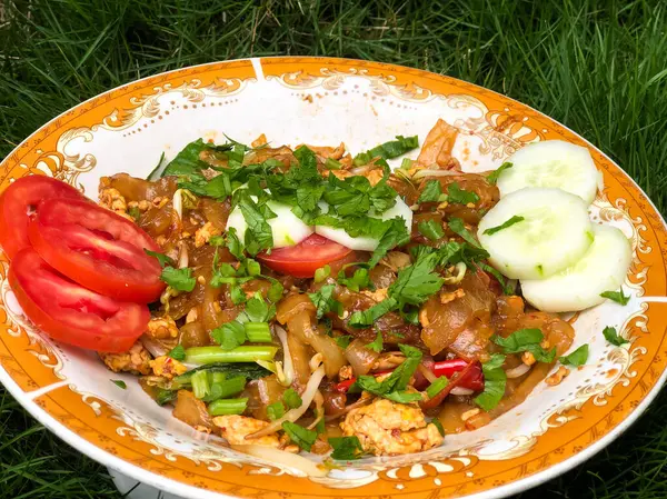 Dilimlenmiş salatalık, domates ve kereviz yaprakları gibi çeşitli garnitürlerle servis edilen bir tabak Tiaw Noodles.