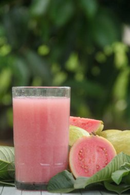 Guava suyu ya da bir bardakta Psidium Guajava ve yeşil yaprakların doğal bir arka planına sahip bir masada tüm ve dilimlenmiş guava ile süslenmiş.