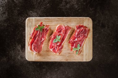 Serrano jambonu ekmek dilimlerinde servis etti. Koyu arkaplandaki tahta kesme tahtasının üst görüntüsü. Tipik İspanyol yemeği..