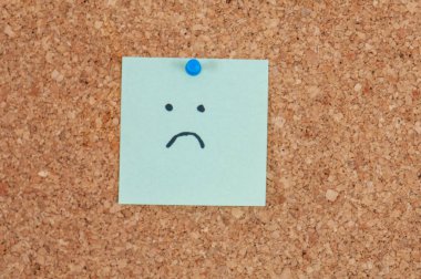 Yakın çekim, üzerinde üzgün bir yüz çizilmiş açık mavi, kare yapışkan bir nota gösterir, mavi bir iğne ile bir mantar panoya bağlanır, kasvetli ve dokunaklı bir görüntü yaratır..