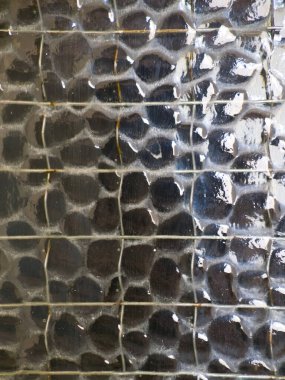 Gömülü bir tel örgü ızgarası ve pürüzlü, çakıllı bir yüzey içeren desenli güçlendirilmiş camların detaylı makro fotoğrafı. Cam yüzey ışığı yansıtır ve soyut bir desen oluşturur..