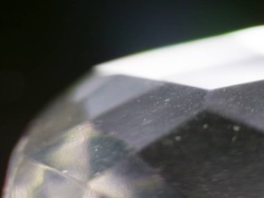 Çok detaylı bir makro fotoğraf şeffaf bir taşın yüzeyini ve kenarlarını yakalıyor. Görüntü, kristalin berraklığını ve dokusunu vurgulayarak karmaşık kesiklere ve ince yansımalara odaklanıyor. Arka plan karanlık.
