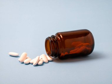 An amber glass pill bottle lies on its side with several white tablets spilled out on a light blue surface. The image highlights medication, pharmacy, and healthcare concepts in a clean, clinical setting.
