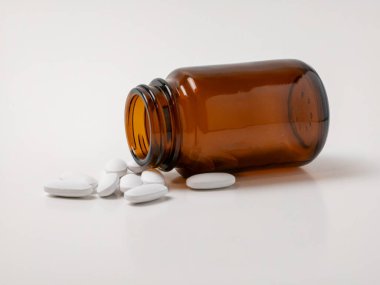 Kahverengi bir cam şişe beyaz bir yüzeye dökülen beyaz yuvarlak haplarla yan yatar. Görüntü, sağlık, ilaç, eczane ve reçeteli ilaçlarla ilgili konuları temiz ve klinik bir ortamda aktarmaktadır..