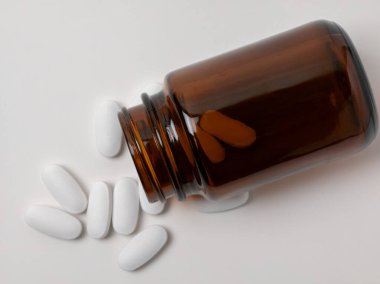 Kahverengi bir cam ilaç şişesi yan yatmış beyaz bir yüzeye yayılmış birkaç beyaz oblong tablet ile birlikte. Görüntü steril bir ortamda sağlık, ilaç ve tedavi kavramlarını iletir.
