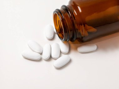 Açık kehribar rengi bir cam şişeden dökülen çok sayıda beyaz oval tablet, ilaç, takviye veya ilaç kavramları izlenimi uyandırarak beyaz bir yüzeye saçılmıştır. Görüntü sağlık, tedavi ve tıbbi konuları iletir