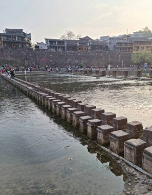 Zhangjiajie, Çin - Nisan 07, 2025: Geleneksel binalar ve turistlerin arka planında yer aldığı nehir boyunca bir yol oluşturan düzenlenmiş kare taşlar serisi