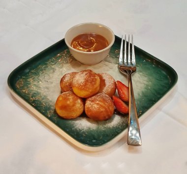 Cakarta, Endonezya - 13 Mayıs 2025: Poffertjes 'in yumuşacık dokusu yeşil bir tabak ve gümüş kaşıkla servis edilen karamel sosu ve dilimlenmiş çilek