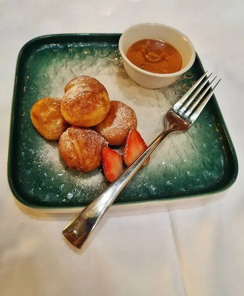 Cakarta, Endonezya - 13 Mayıs 2025: Poffertjes 'in yumuşacık dokusu yeşil bir tabak ve gümüş kaşıkla servis edilen karamel sosu ve dilimlenmiş çilek