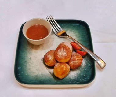 Cakarta, Endonezya - 13 Mayıs 2025: Poffertjes 'in yumuşacık dokusu yeşil bir tabak ve gümüş kaşıkla servis edilen karamel sosu ve dilimlenmiş çilek