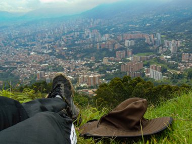 Pantolonu, siyah tenis ayakkabısı, kahve ve su ısıtıcısı olan siyah bir sırt çantası, çimlerin üzerinde oturan ve şehre bakan biri..