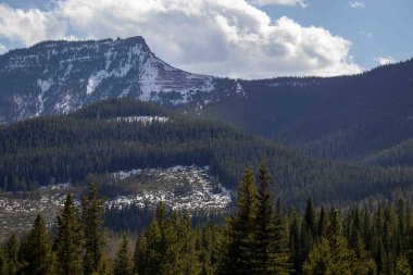 Beyaz bulutlu mavi gökyüzü ve arka planda yükselen yüksek dağlar, etrafa saçılmış yeşil çam ağaçları..