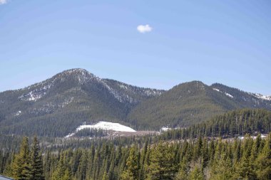Beyaz bulutlu mavi gökyüzü ve arka planda yükselen yüksek dağlar, etrafa saçılmış yeşil çam ağaçları..