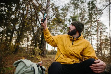 Sakallı bir adam ormanın ortasındaki devrilmiş bir ağacın üzerinde dinleniyor. Adamın biri GPS 'i aramak için cep telefonu kullanıyor..