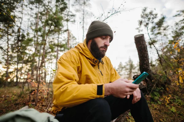一个长胡子的男人躺在森林中央一棵倒下的树上 男人用手机搜索Gps — 图库照片
