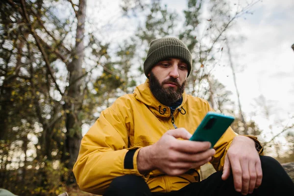 髭を生やした男が森の真ん中の倒木に腰を下ろしている 男はGpsを検索するために携帯電話を使用します — ストック写真