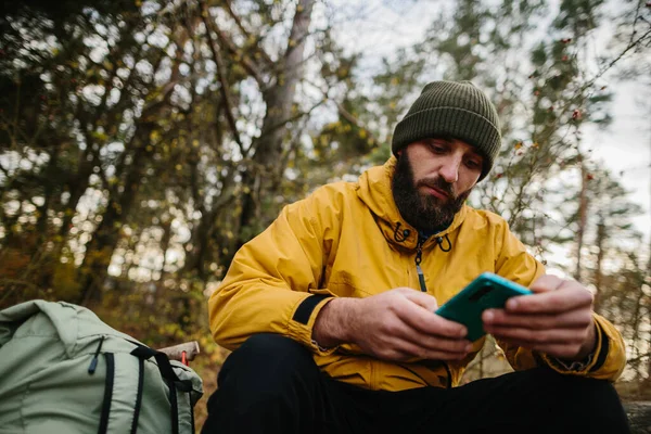 髭を生やした男が森の真ん中の倒木に腰を下ろしている 男はGpsを検索するために携帯電話を使用します — ストック写真