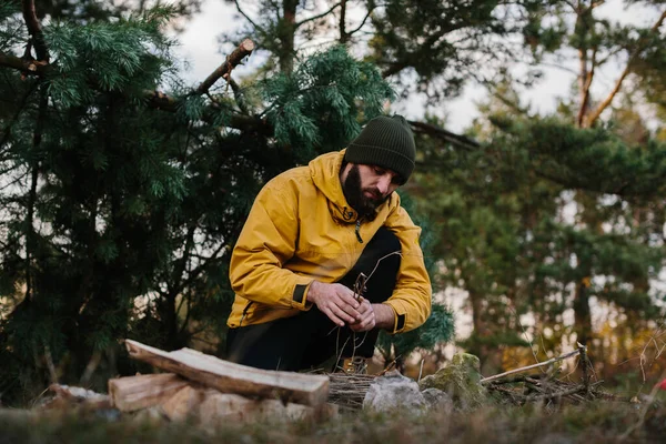在野外生存 一个满脸胡须的男人在一个用松树树枝搭成的临时掩蔽所附近点火 — 图库照片