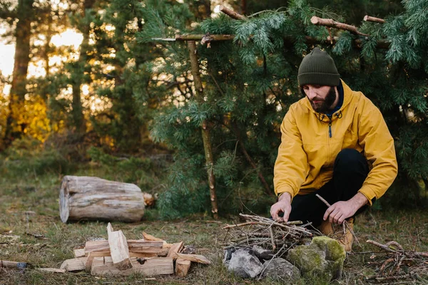 野生での生存 髭を生やした男が松の枝で作られた仮設の避難所の近くの火を灯す — ストック写真