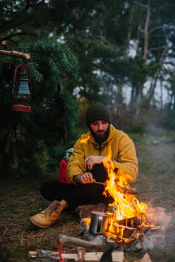 Sakallı erkek otostopçu şenlik ateşinde sosis kızartıyor. Vahşi doğada akşam yemeği, seyahat ve hayatta kalma konsepti.