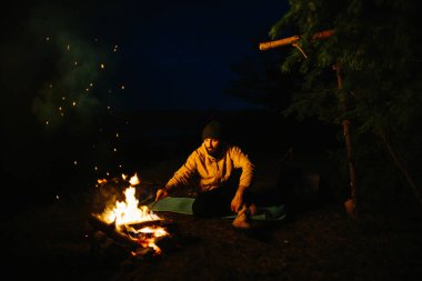 Gezgin gece kamp ateşinin yanında dinleniyor. Gaz lambası ve taşlarla çevrili bir ateşle uyumak için dallardan korunaklı bir barınak..