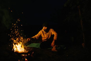 Gezgin gece kamp ateşinin yanında dinleniyor. Gaz lambası ve taşlarla çevrili bir ateşle uyumak için dallardan korunaklı bir barınak..