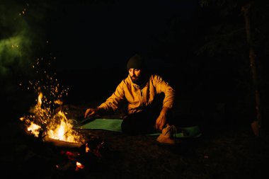 Gezgin gece kamp ateşinin yanında dinleniyor. Gaz lambası ve taşlarla çevrili bir ateşle uyumak için dallardan korunaklı bir barınak..