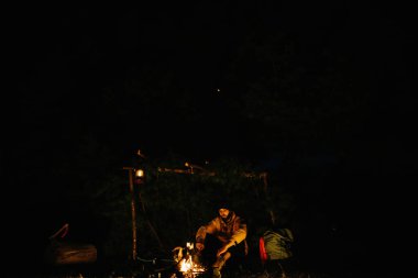 Gezgin gece kamp ateşinin yanında dinleniyor. Gaz lambası ve taşlarla çevrili bir ateşle uyumak için dallardan korunaklı bir barınak..