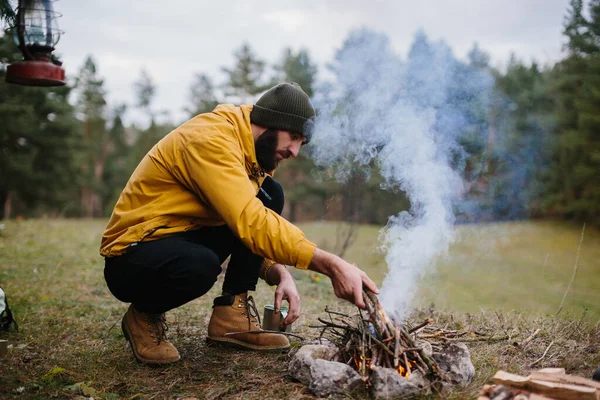 野生での生存 髭を生やした男が松の枝で作られた仮設の避難所の近くの火を灯す — ストック写真