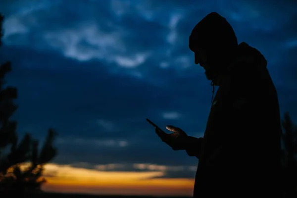 A man stands with a phone against the background of the night sky. Hiker in the mountains at sunset.