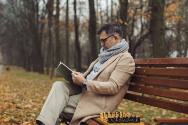 Yaşlı bir adam sonbahar parkında bir bankta otururken dergi okur. Bir adam arkadaşının satranç oynamasını bekliyor..