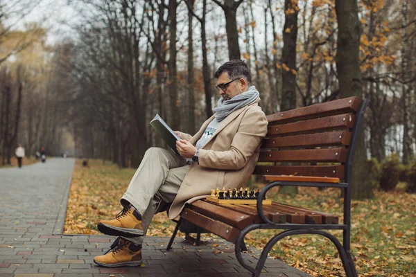 秋の公園のベンチに座っていると 老人が雑誌を読んでいる 男は友達がチェスをするのを待っている — ストック写真