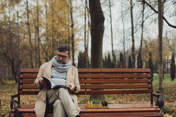 Yaşlı Bir Adam Sonbahar Parkında Bir Bankta Otururken Dergi Okur — Stok fotoğraf