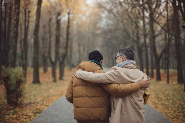 Friends, a senior and a young man walking and talking and drinking coffee together in the autumn park