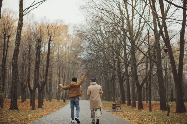 先輩と若い男が一緒に秋の公園でコーヒーを飲みながら歩いています — ストック写真